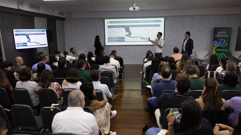 Foto de pessoas na plateia com o slide e os palestrantes.