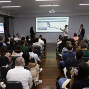 Foto de pessoas na plateia com o slide e os palestrantes.
